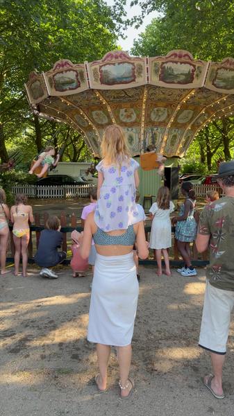 A woman holds a child on her shoulders as they watch a colorful carousel ride in an outdoor amusement park on a sunny afternoon